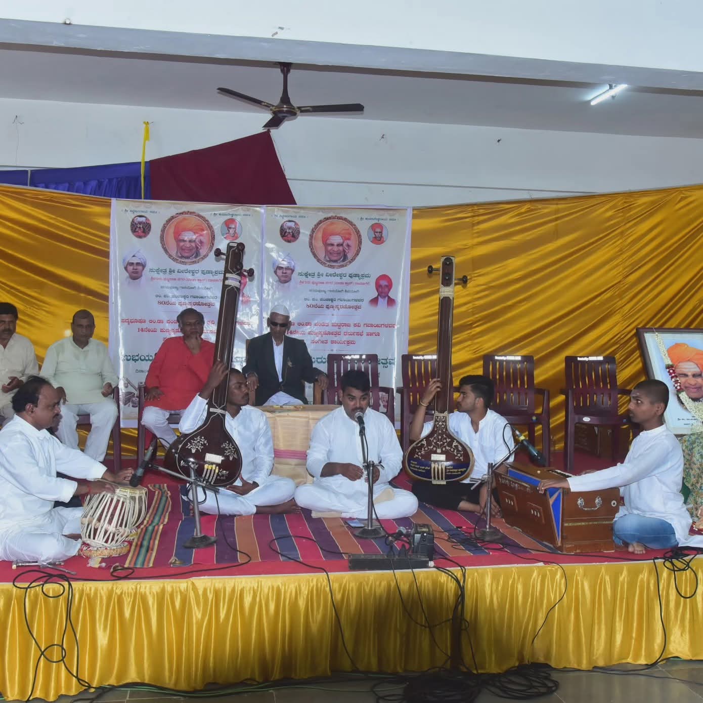 Students and teachers performing together on stage with tabla, tanpura and harmonium
